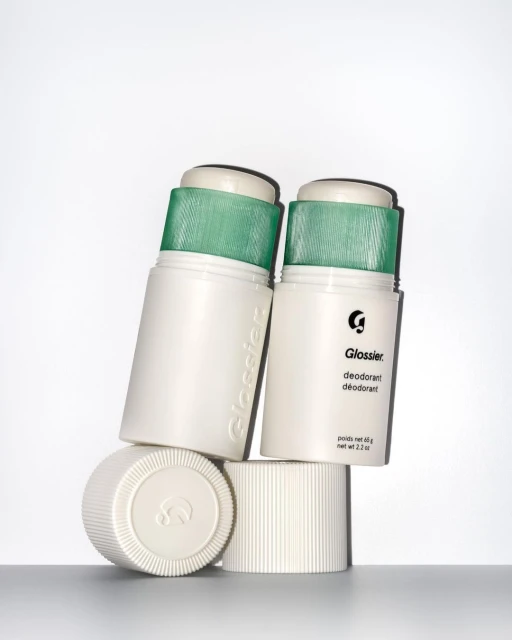 Close-up studio shot of two white Glossier deodorant sticks with green caps, one leaning against the other, and two white caps.