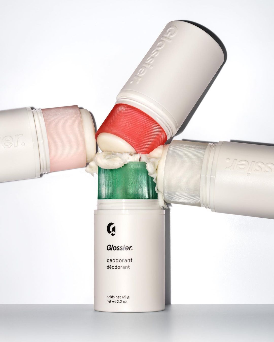 Four Glossier deodorant sticks, each with a different colored applicator, rest on top of one another against a white backdrop in a studio.