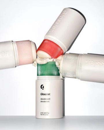 Four Glossier deodorant sticks, each with a different colored applicator, rest on top of one another against a white backdrop in a studio.