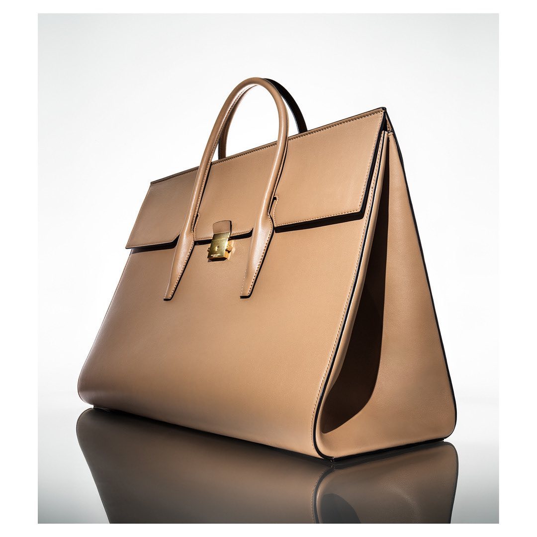 Tan leather handbag with gold hardware displayed on a reflective white studio surface by Raymond Meier