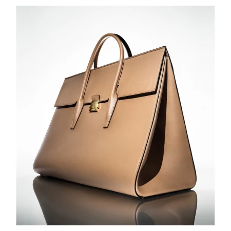 Tan leather handbag with gold hardware displayed on a reflective white studio surface by Raymond Meier