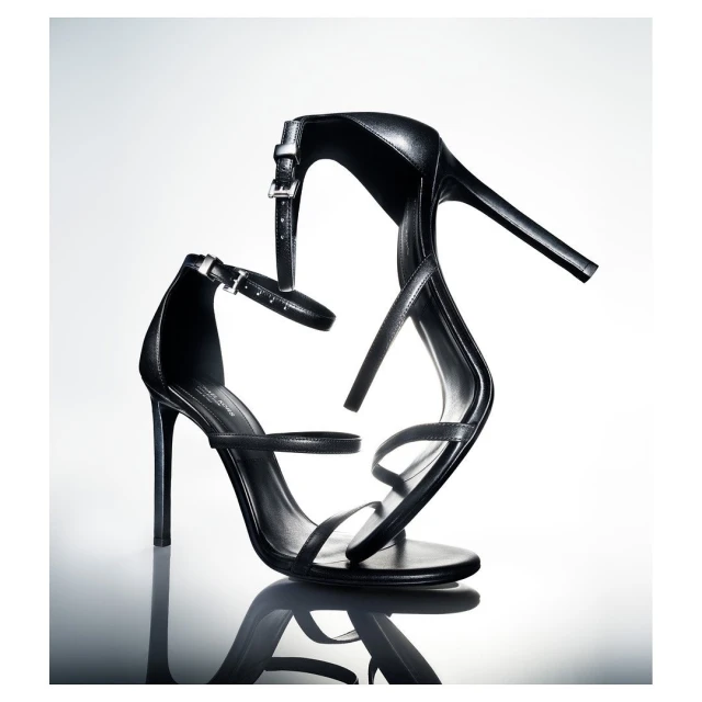 Black leather stiletto heeled sandals are reflected on a glossy surface against a bright gradient white background in a studio shot.