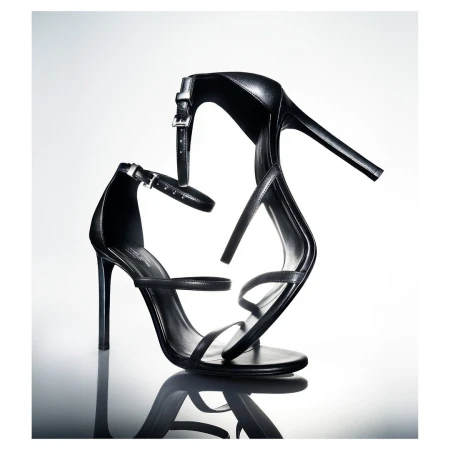 Black leather stiletto heeled sandals are reflected on a glossy surface against a bright gradient white background in a studio shot.
