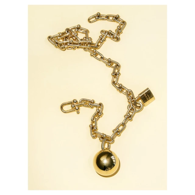 Gold chain necklace with padlock and ball pendant on a light yellow backdrop, top-down view. Jewelry photography.