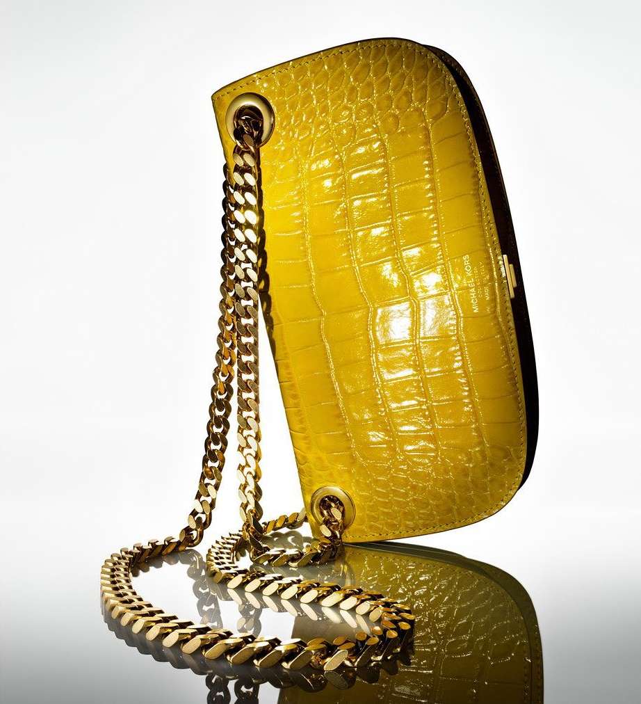Yellow croc-skin handbag with chunky gold chain strap on a reflective surface, illuminated by soft lighting against a bright white backdrop.