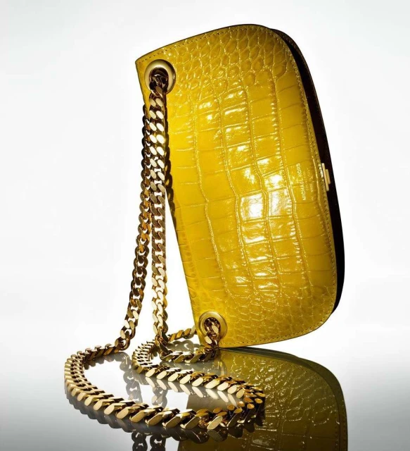 Yellow croc-skin handbag with chunky gold chain strap on a reflective surface, illuminated by soft lighting against a bright white backdrop.