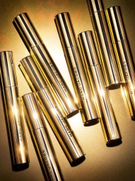 Multiple metallic gold Marc Jacobs cosmetic tubes arranged diagonally against a warm golden gradient background.