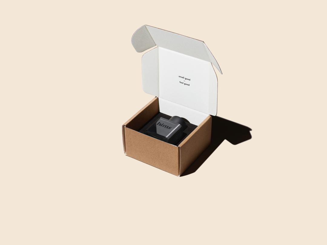 Open cardboard box on a beige background. Inside the box is a dark grey product with white text on top; the box is lined with white.