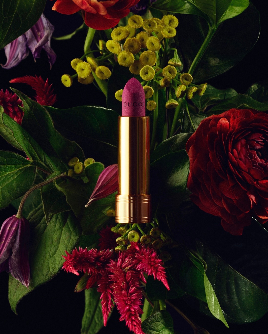 Close-up shot of Gucci lipstick surrounded by vibrant assortment of flowers including roses and celosia on a black background.