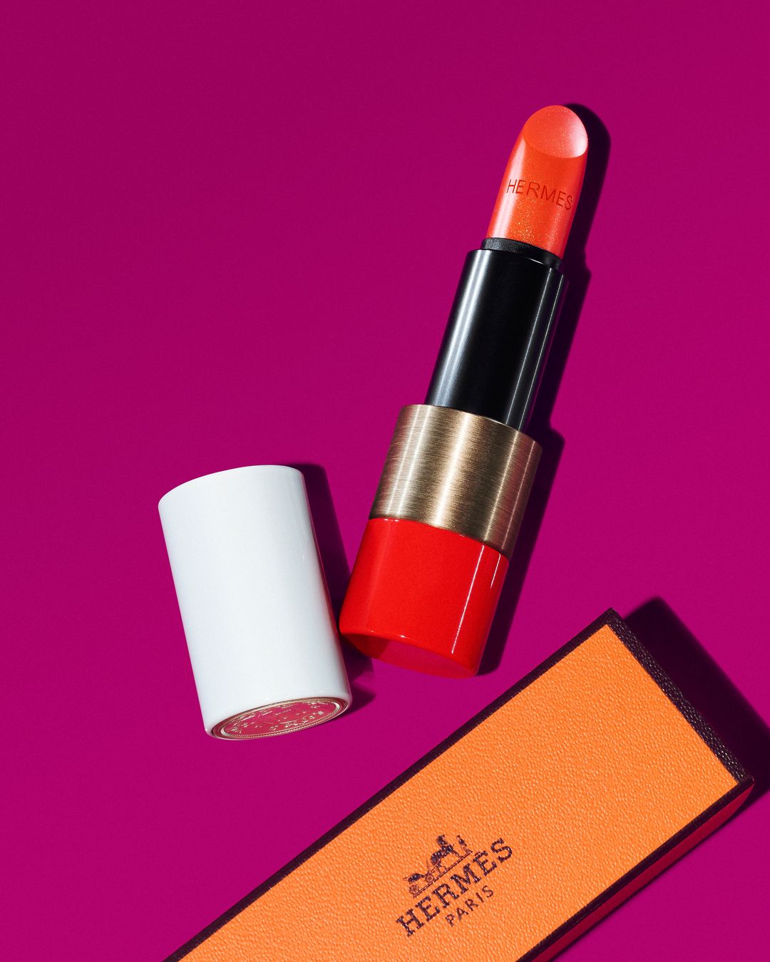 Studio shot of a vibrant orange Hermes lipstick with its cap and branded box against a magenta background, product focus, high-end beauty.