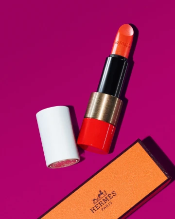 Studio shot of a vibrant orange Hermes lipstick with its cap and branded box against a magenta background, product focus, high-end beauty.