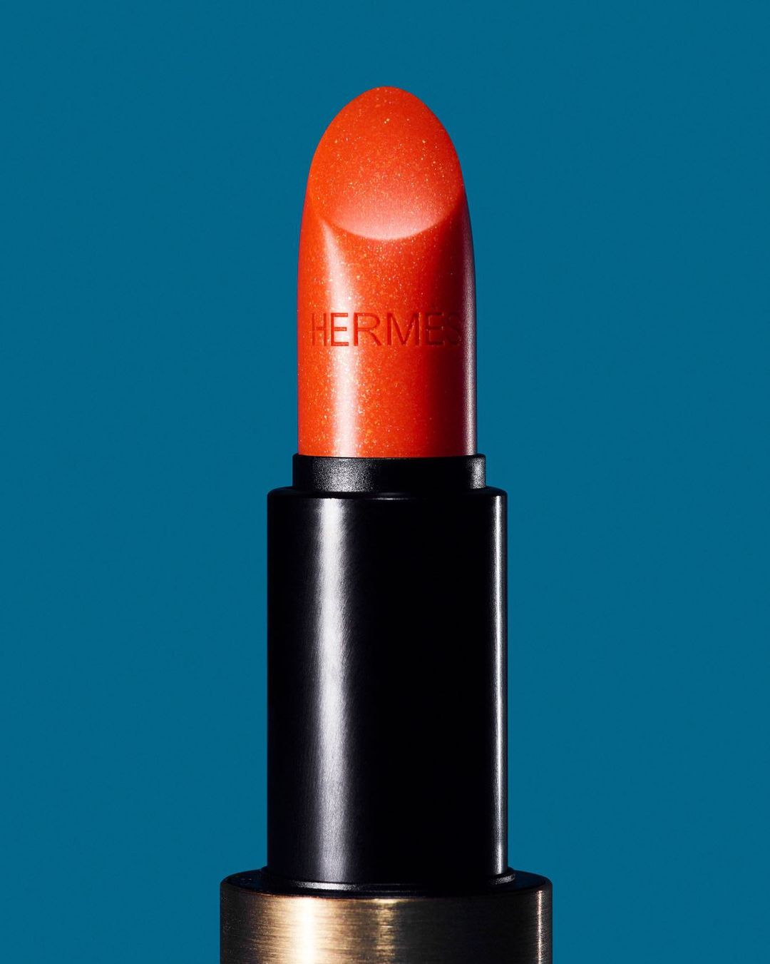 Close-up of a vibrant orange Hermès lipstick, showcasing its sleek black and gold packaging against a teal background.