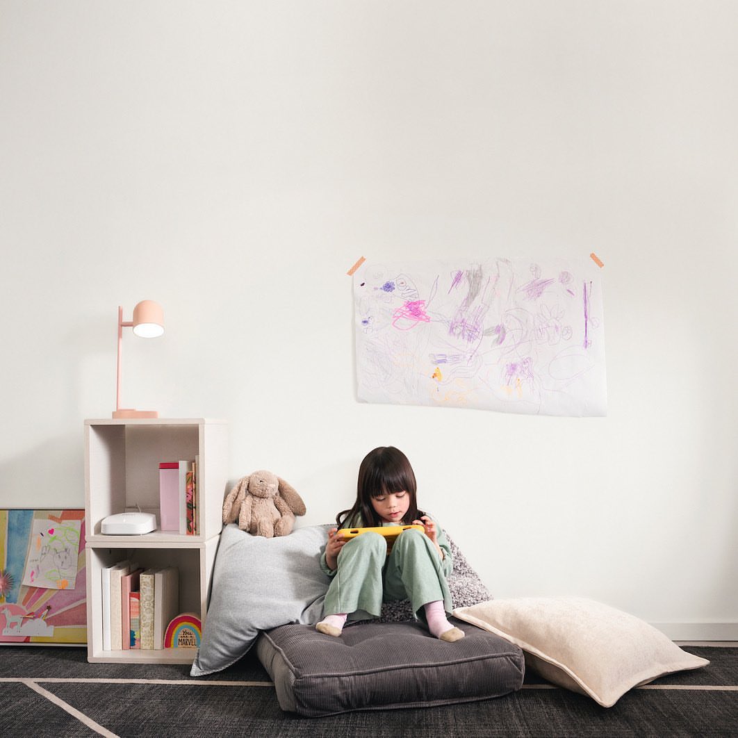 A child sits on a gray floor cushion in an indoor setting playing a handheld video game next to a low bookshelf and children's art.