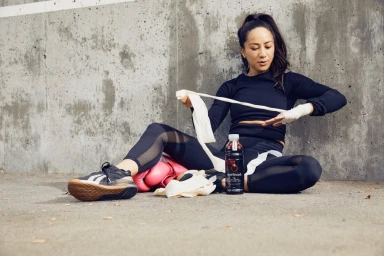 Woman in black athletic wear wrapping her hands with white tape while seated against a concrete wall