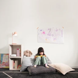 Young child seated on floor pillows playing with a handheld device in a brightly lit room with artwork taped to the wall.