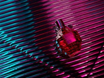 Faceted glass perfume bottle resting on a corrugated metallic surface with vibrant pink and cyan lighting.