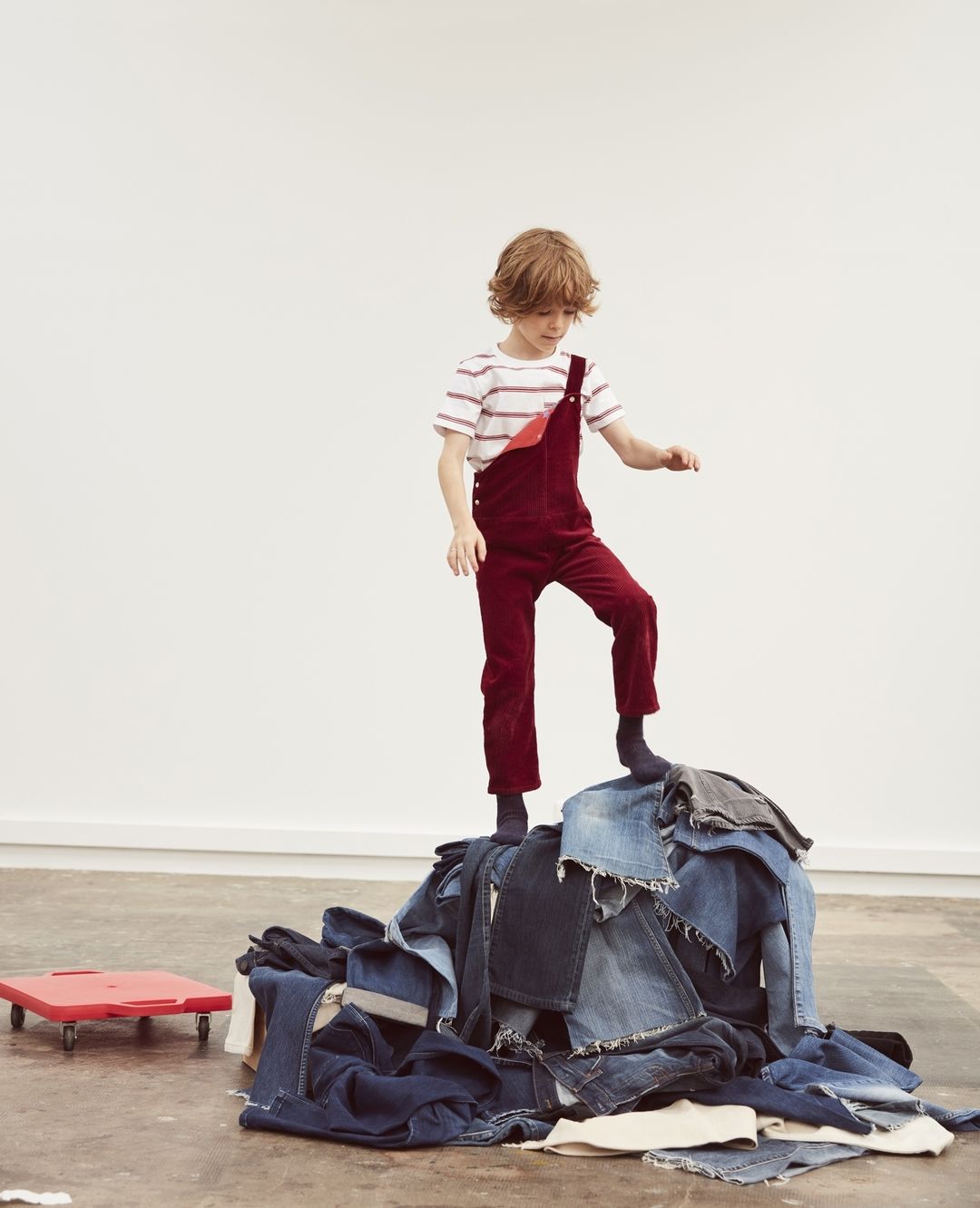 Boy stands atop a pile of denim jeans in a light photo studio. He wears a striped shirt and red overalls, bright indoor lighting.