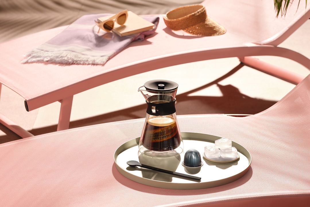 Coffee carafe and pod on a tray, styled with sunglasses and sunhat on pink lounge chairs, bathed in soft light and shadow.