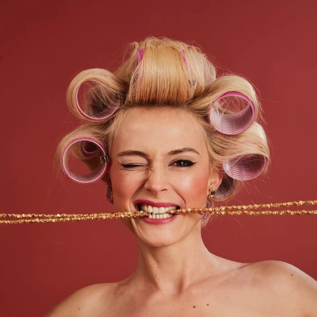 Portrait of a blonde woman with curlers, gold necklace in her mouth, and winking against a red background in a beauty shot.