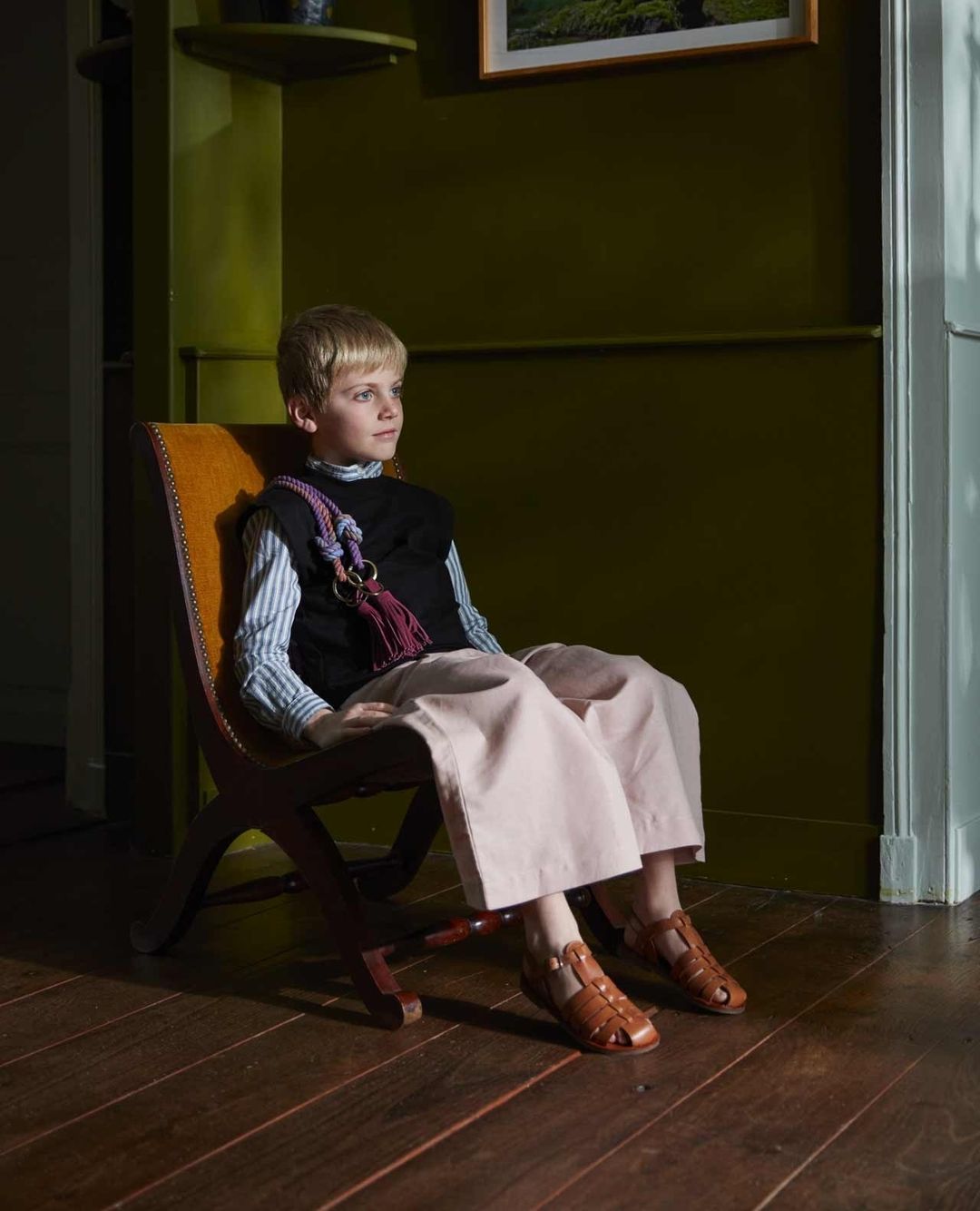 Young boy sits in a wooden chair wearing wide legged pants and sandals, with a striped shirt and black vest in an olive green room.