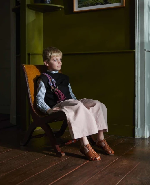 Young boy sits in a wooden chair wearing wide legged pants and sandals, with a striped shirt and black vest in an olive green room.