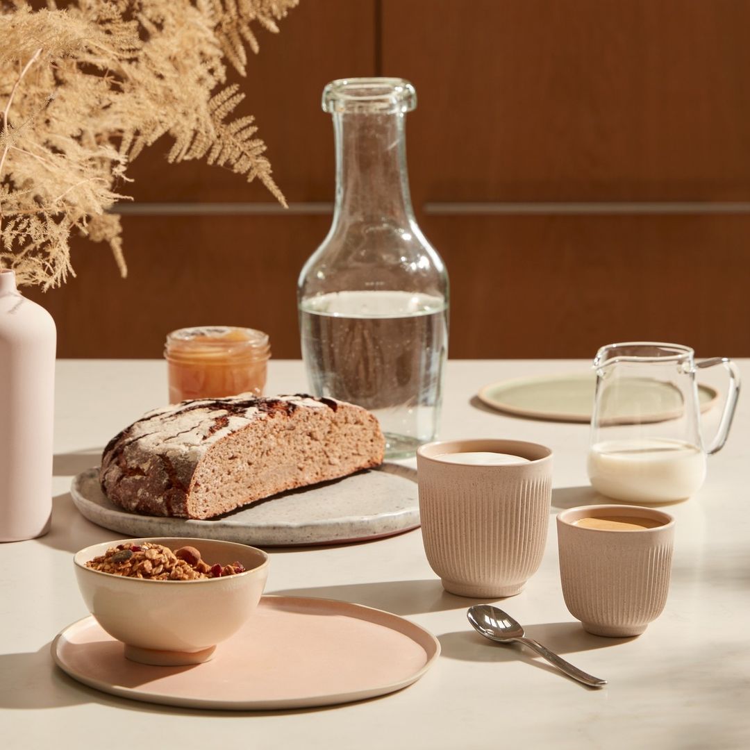 Still life of healthy breakfast on a table featuring bread, cereal, beverages, and fruit under soft, natural lighting.