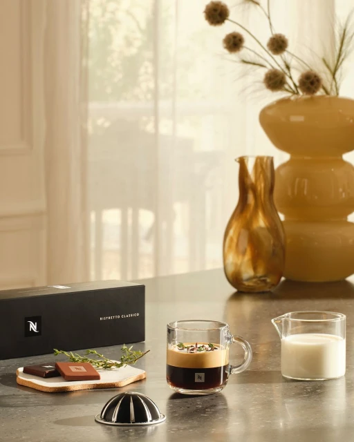 Still life shot of Nespresso box, coffee cup, chocolates, milk and flowers on a counter top in a bright, naturally lit room.