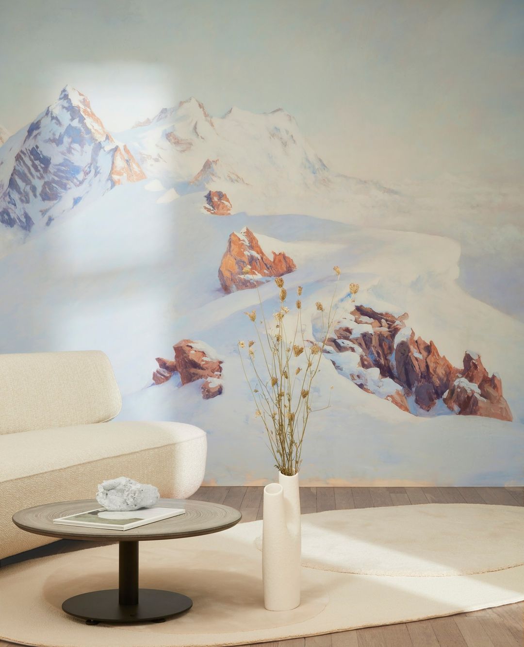 Living room interior with a cream sofa, round table with decor, a white vase with dried flowers, and a mountain mural backdrop.