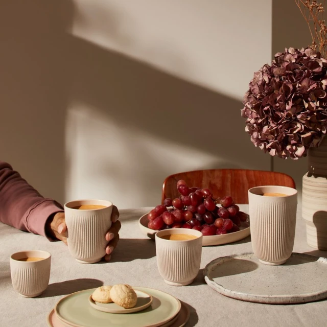 Still life featuring cups of beverage, grapes, cookies and plates arranged on a tablecloth, showcasing a cozy and inviting scene.