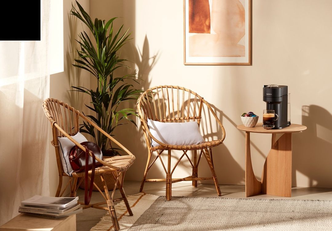Wicker chairs and coffee machine in bright neutral interior with potted plant, artwork, on rug next to round wood accent table.