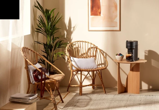 Wicker chairs and coffee machine in bright neutral interior with potted plant, artwork, on rug next to round wood accent table.