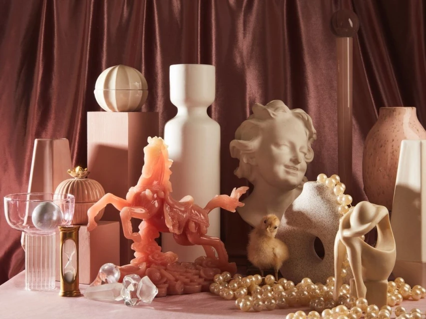 Still life featuring pink horse sculpture, white vase, and bust on pink tablecloth against maroon curtain backdrop with elegant composition.
