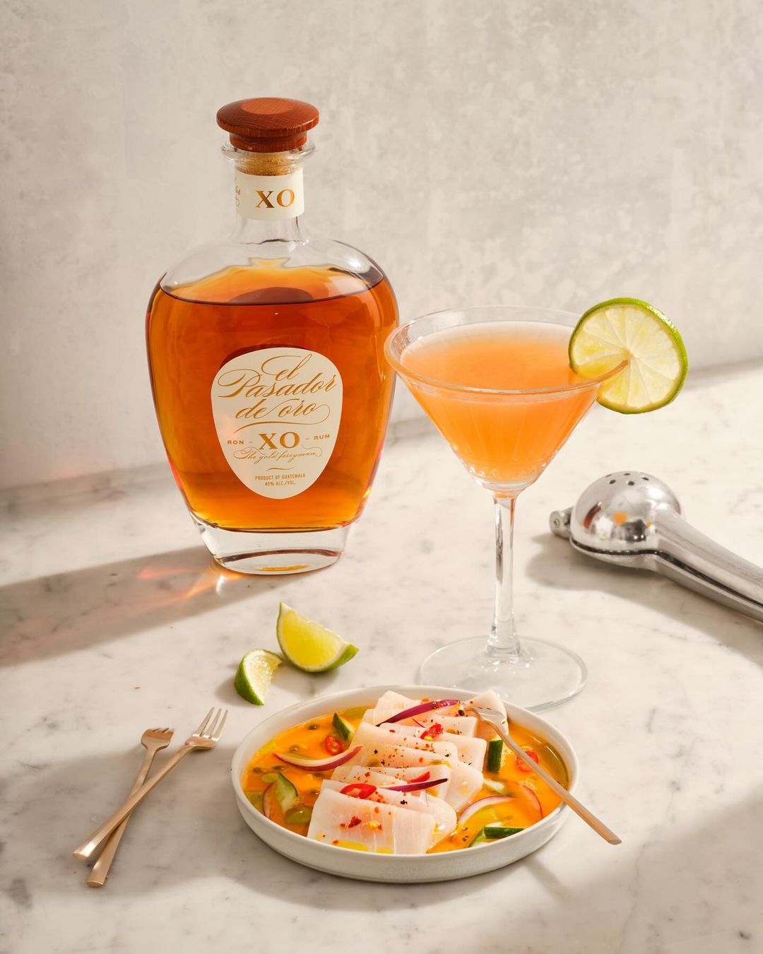 Bottle of El Pasador de Oro XO rum with martini, ceviche appetizer, and cocktail shaker, on a marble surface with neutral background.