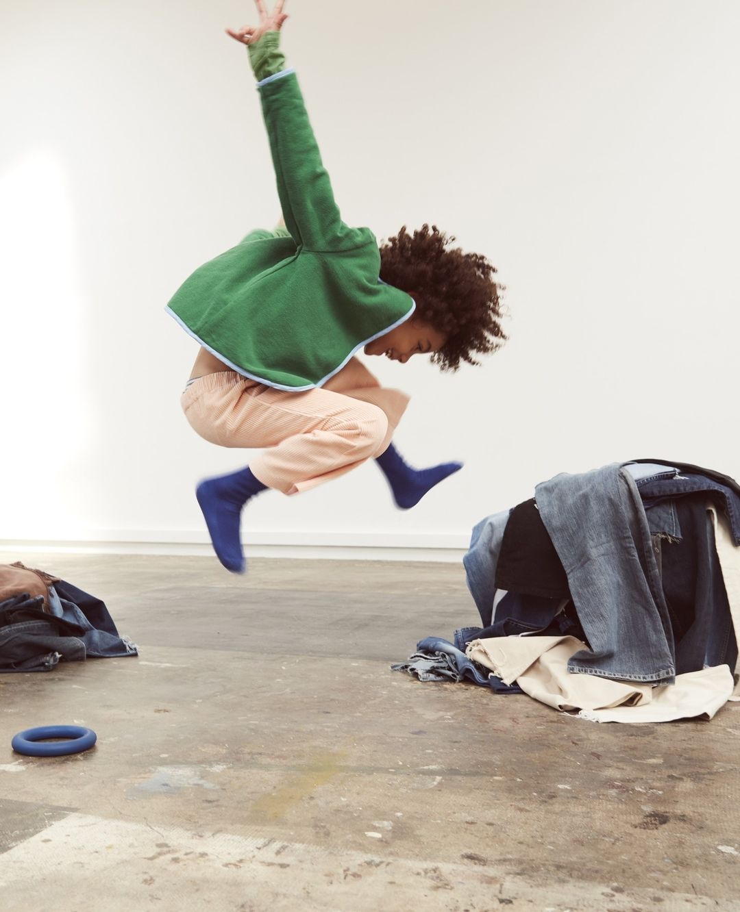 Boy leaping in air indoors with pile of jeans and blue ring toy. Green sweatshirt, peach pants, blue socks, and white wall backdrop.