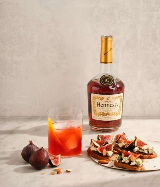 Still life shot of Hennessy cognac bottle, cocktail in a glass with ice, and fig and cheese crostini on a marble surface.
