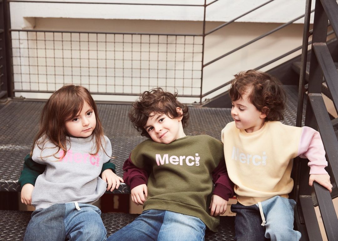 Three cute children pose on metal stairs wearing color-blocked sweatshirts with 'Merci' text, styled with jeans in a casual indoor setting.