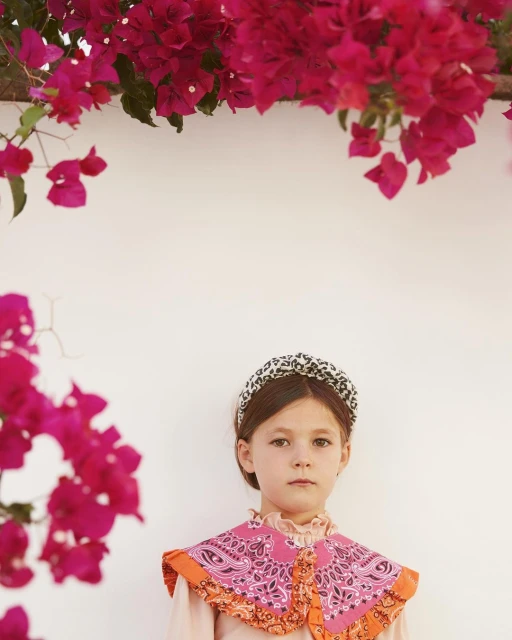 Portrait of a girl wearing a patterned headband and pink bandana collar, standing in front of a white wall with bougainvillea flowers.