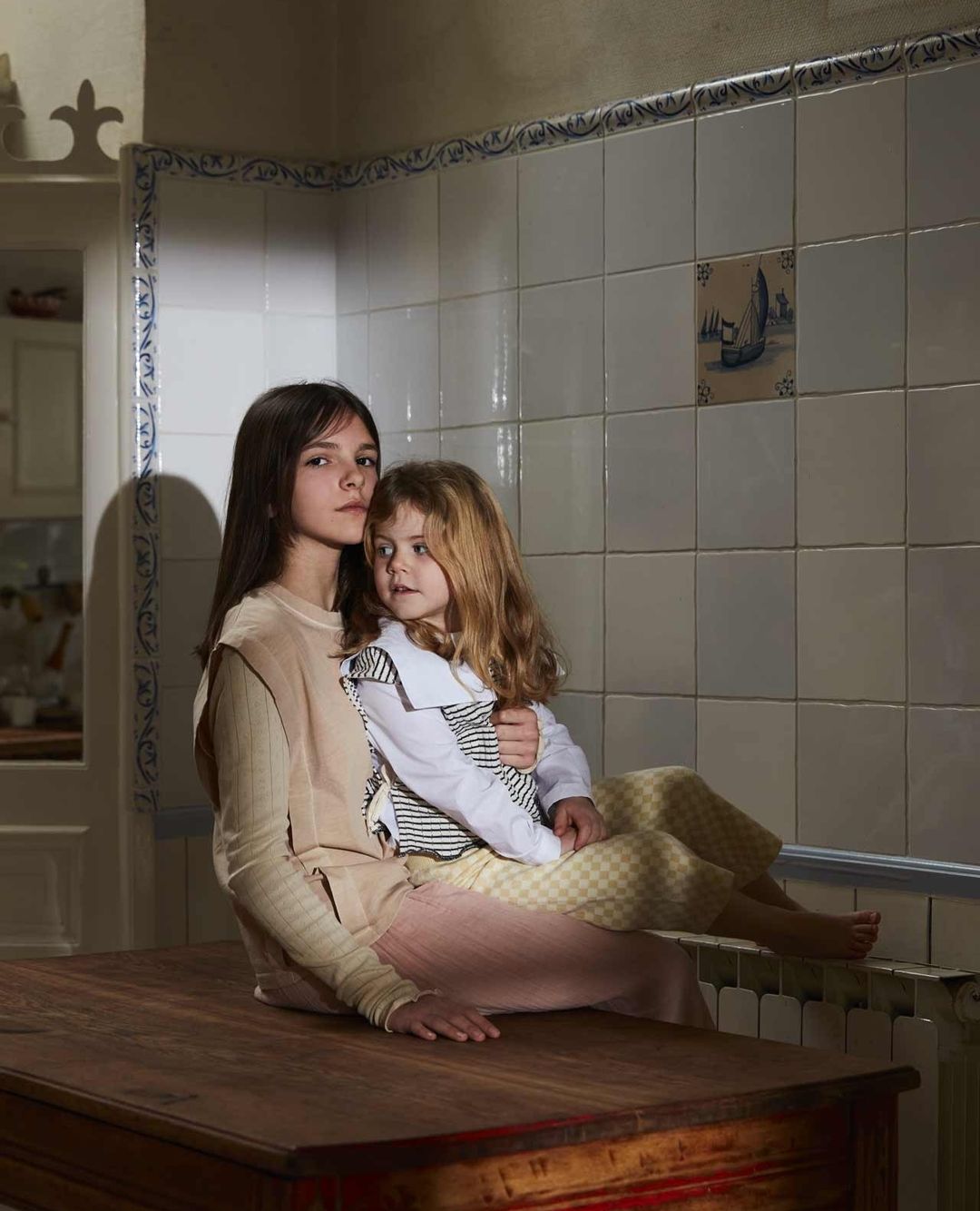 A girl holds a toddler while sitting on a wooden table in an indoor setting with tiled walls and neutral lighting. The pair is centered.
