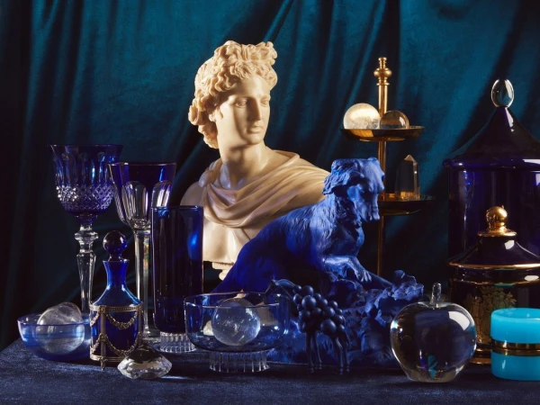 Still life with bust, dog statue, glassware and dark-blue velvet backdrop. Features crystal, glass, and marble material elements.