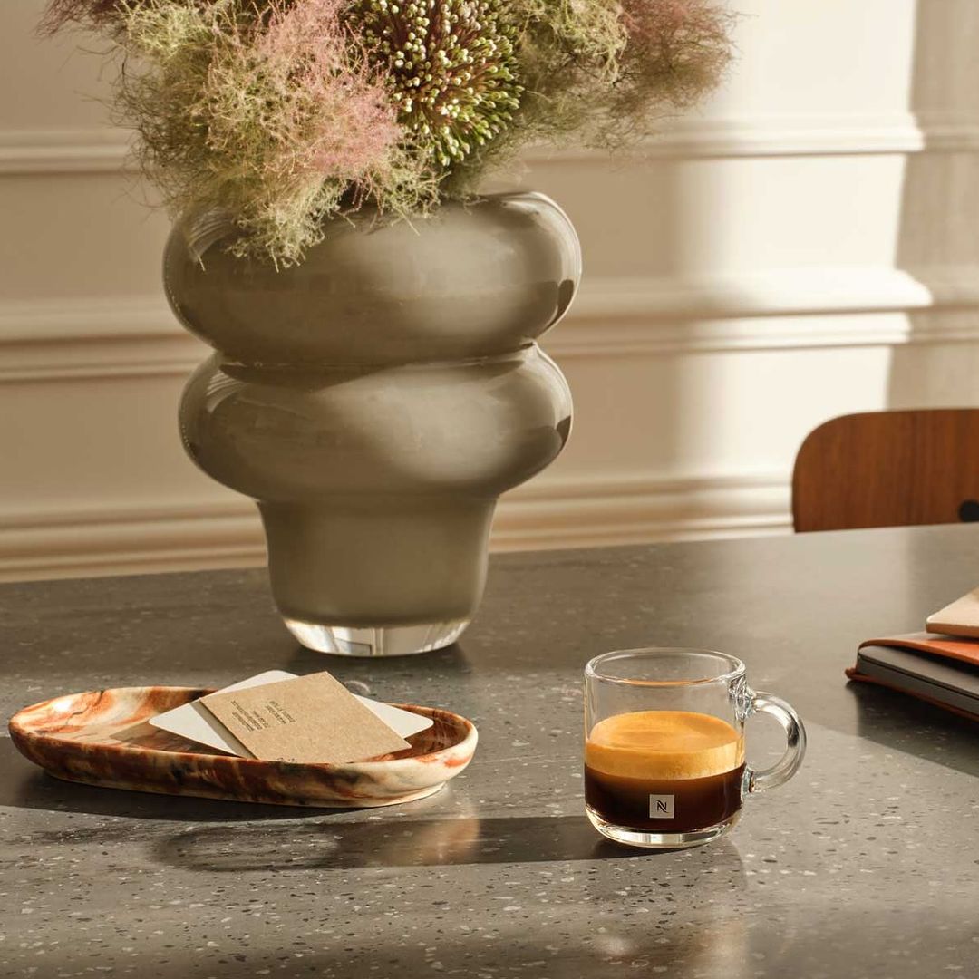 Still life features a glass mug of espresso, a vase with flowers, and a marble tray on a gray table in a naturally lit room.