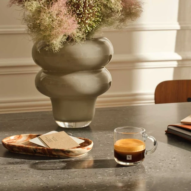 Still life features a glass mug of espresso, a vase with flowers, and a marble tray on a gray table in a naturally lit room.