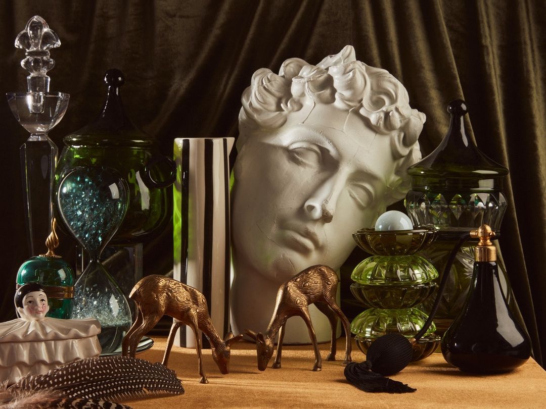 Ornate still life with glass jars, sculpture head, deer figurines, and feathers against a draped velvet background in dark, muted tones.