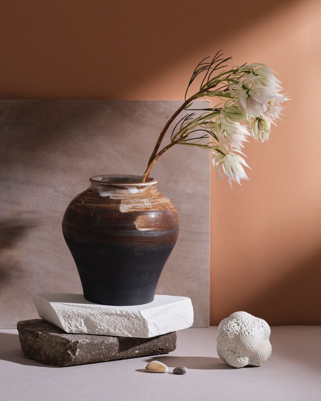Still life composition featuring a vase holding white flowers, arranged on stacked stones against a warm-toned backdrop.