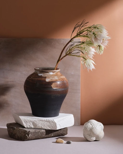 Still life composition featuring a vase holding white flowers, arranged on stacked stones against a warm-toned backdrop.