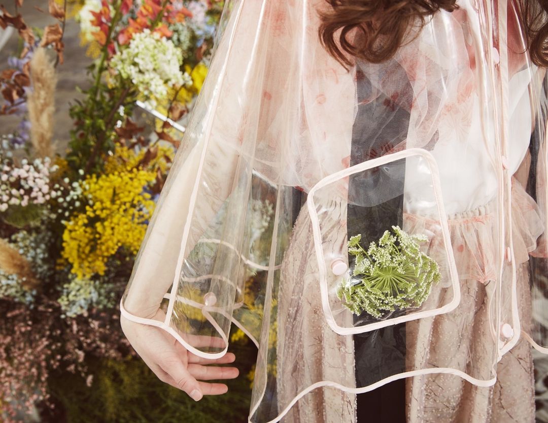 Woman wearing a transparent pink raincoat with a clear pocket filled with green and white flowers, standing in front of flower wall.