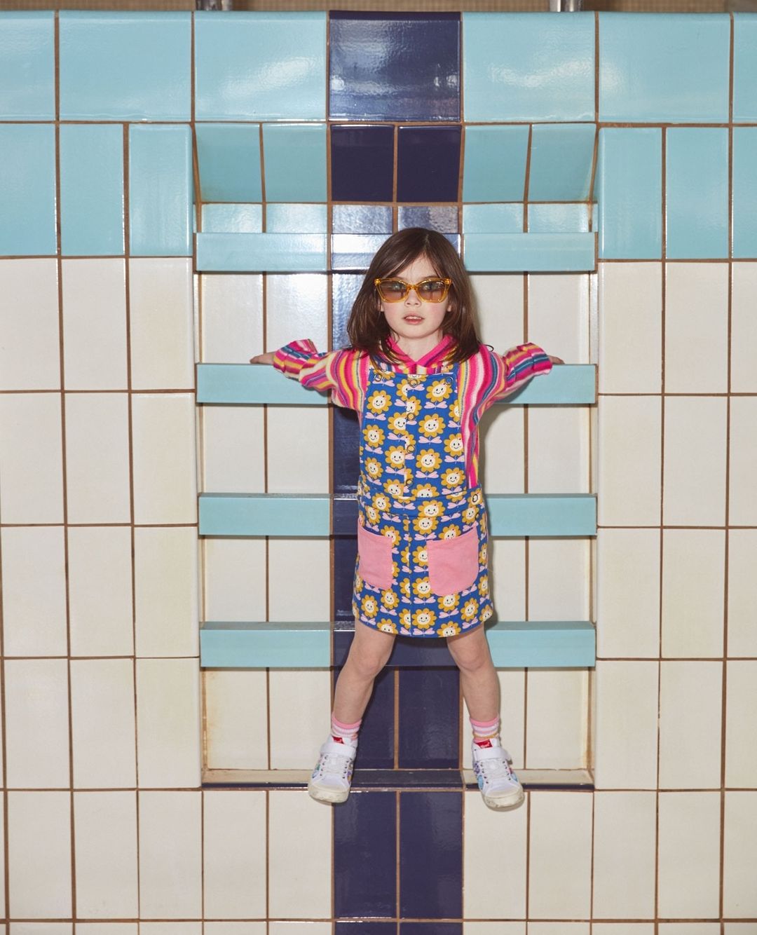 A girl in sunglasses and patterned dress leans against a colorful tiled wall, posing in a unique, eye-level studio shot.