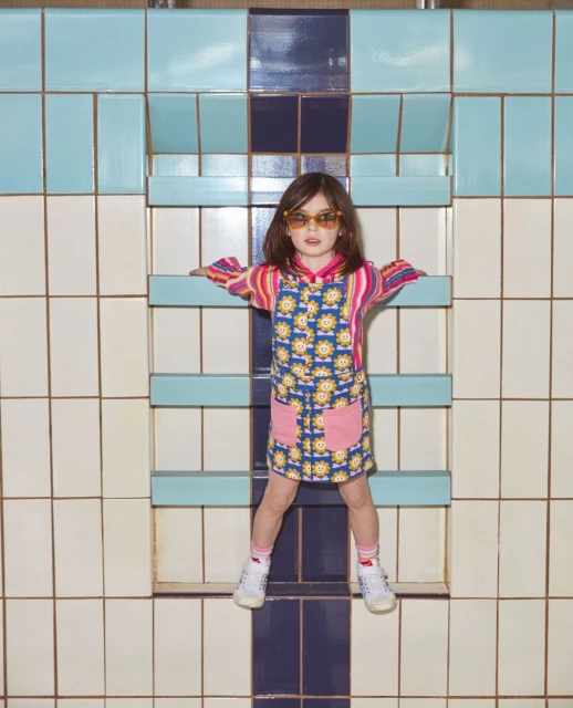 A girl in sunglasses and patterned dress leans against a colorful tiled wall, posing in a unique, eye-level studio shot.