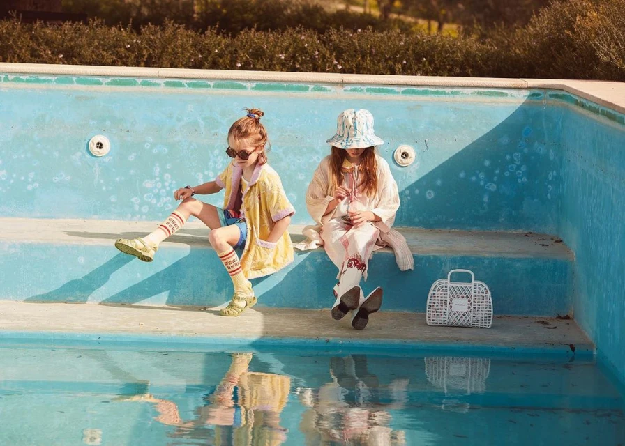 Two young girls in stylish clothing sit on the edge of an empty swimming pool on a sunny day, enjoying the outdoor setting.