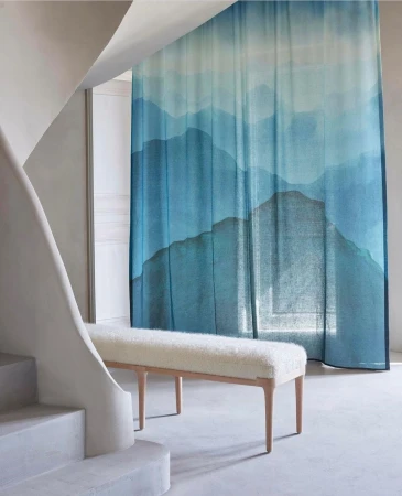 Interior design featuring a long cream-colored bench with wooden legs in front of a gradient blue mountain-themed curtain, near white stairs.