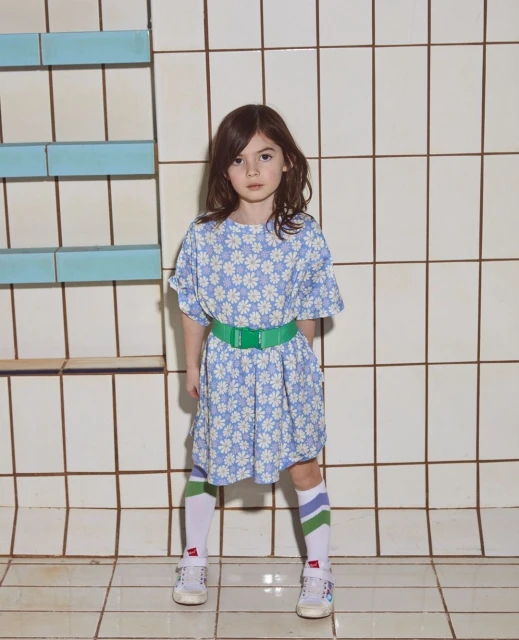 A girl in a floral dress stands in front of a tiled wall, wearing striped socks and white sneakers, casting a shadow to her right.